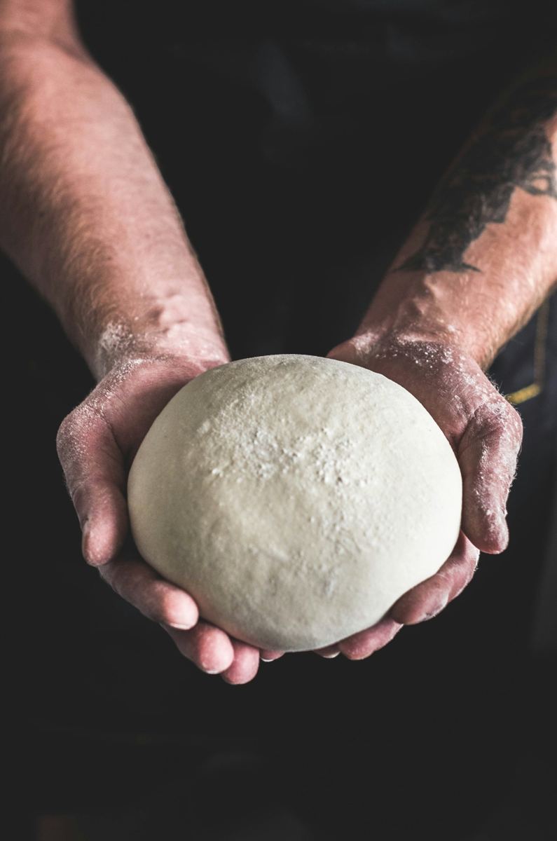 A baker holding a round of proved dough in both hands against a dark background.