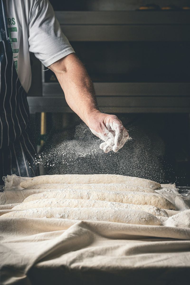 A baker dusting flour over dough on the bench.