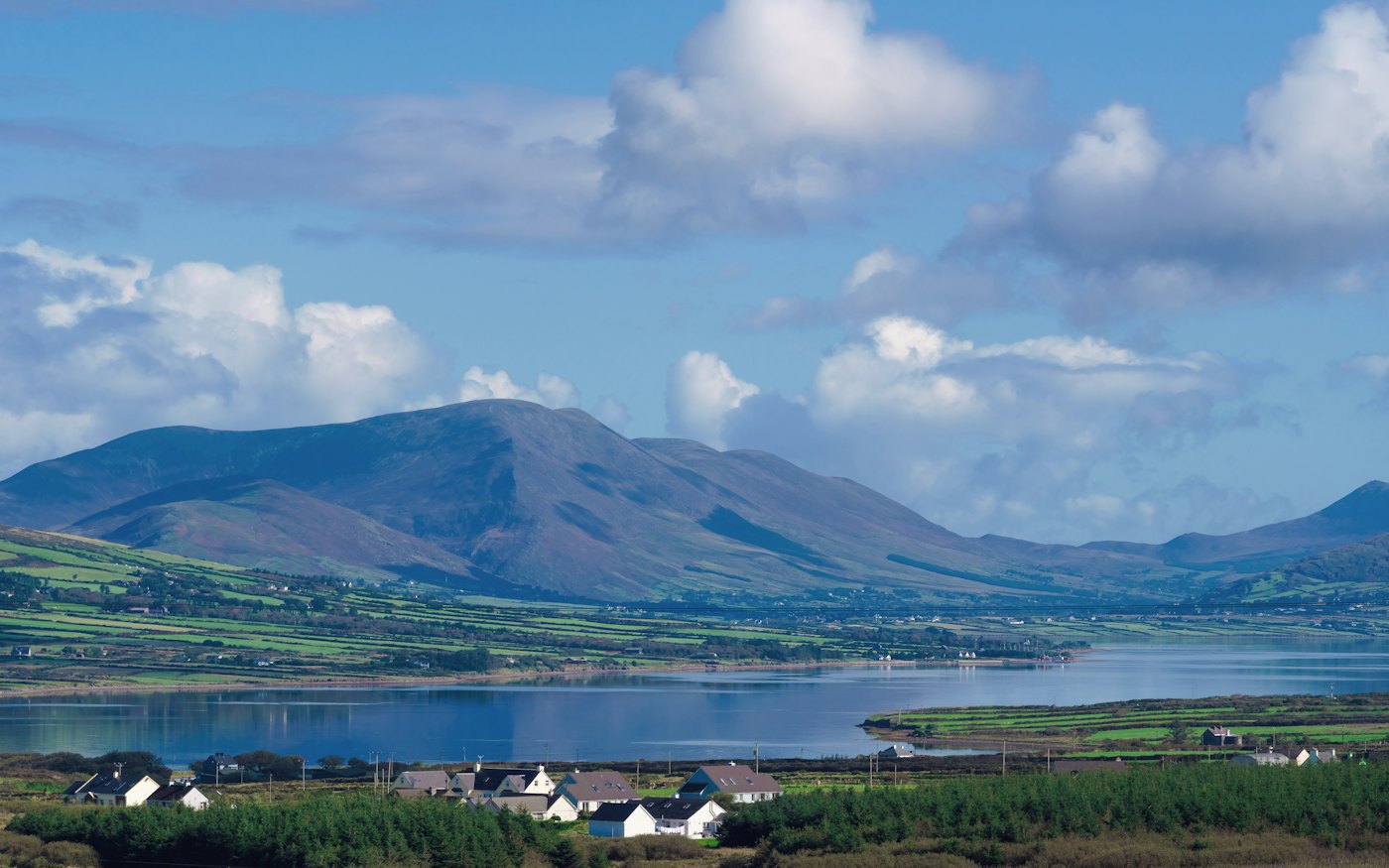A grassy shore and lake beneath mountains, with small white houses in the distance.
