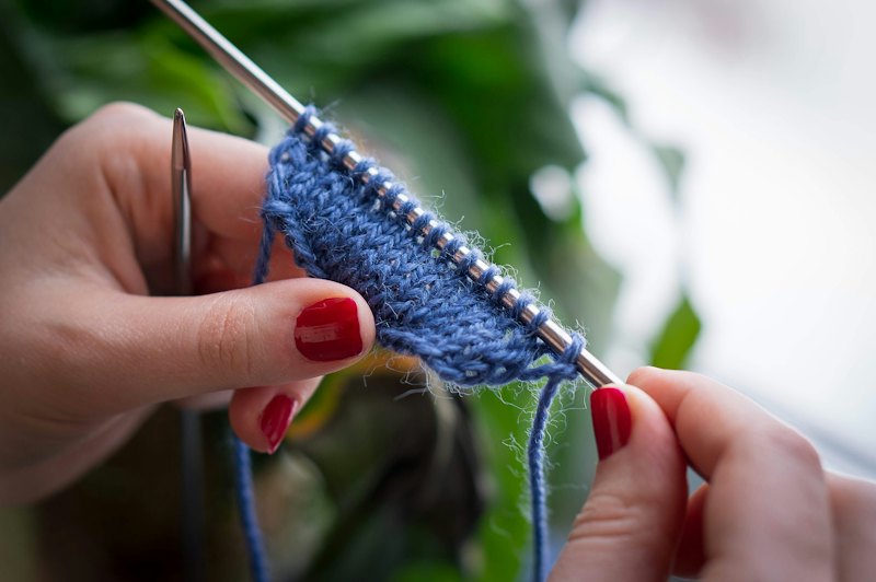 Hands knitting with blue wool on fine needles.