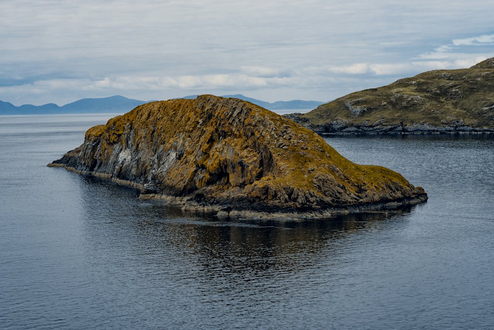 A small island in the sea with clouds and distant hills behind it.