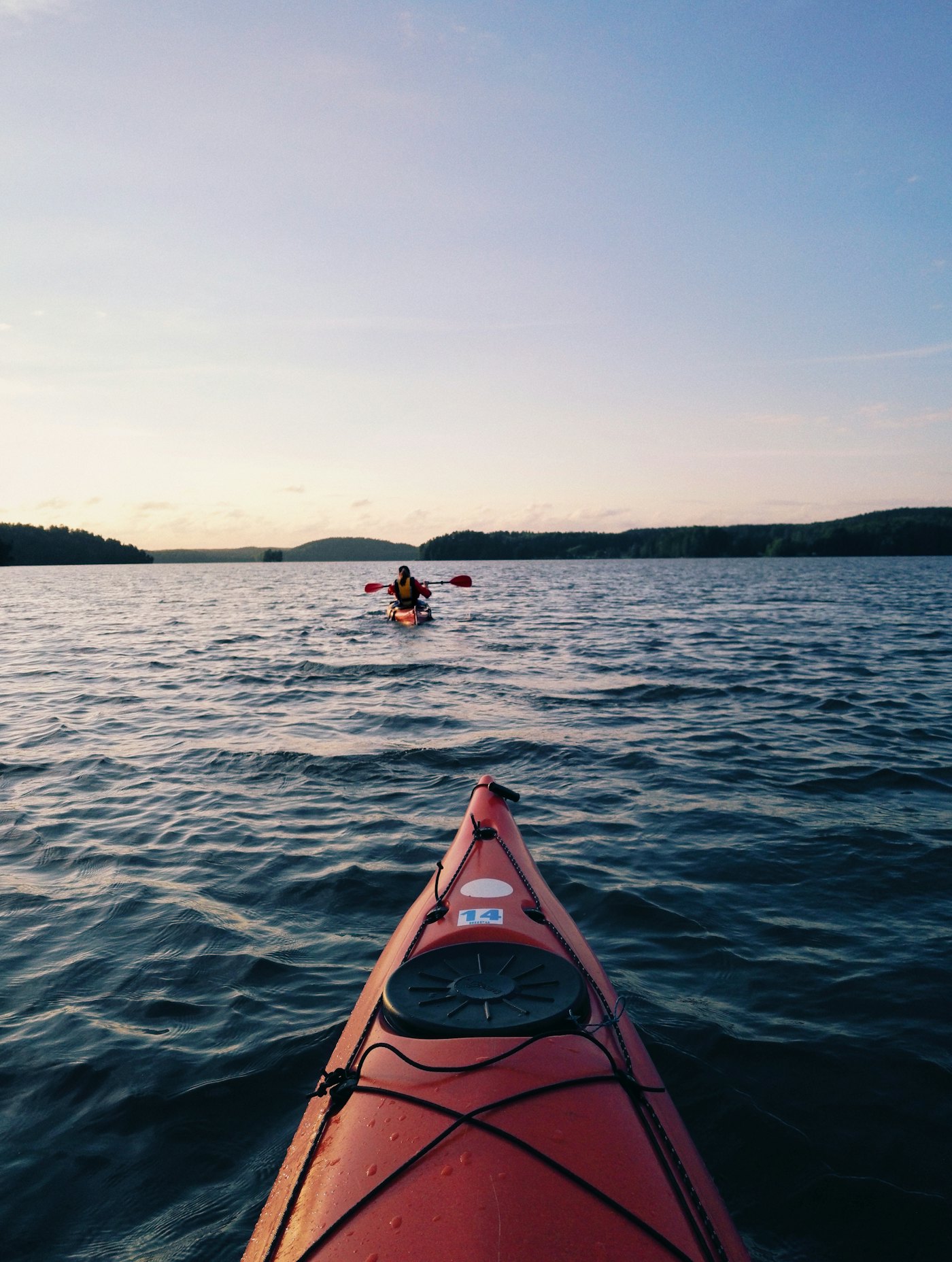 The bow of a red kayak on calm water, with a dusky shoreline in the distance.