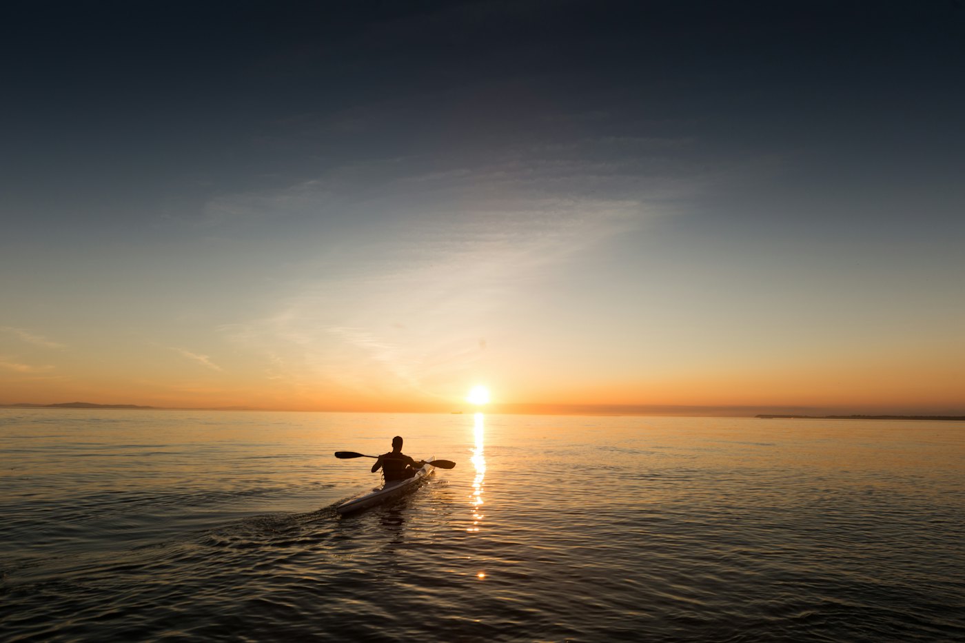 A paddler in silhouette on orange water in front of the setting sun.